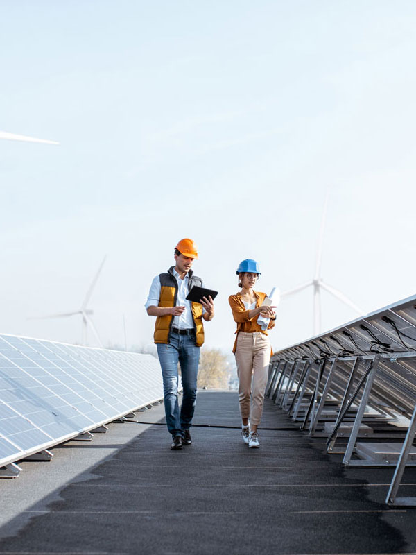 Two workers in orange high-visibility vests stand between rows of PV solar modules and wind turbines on a sunny day.