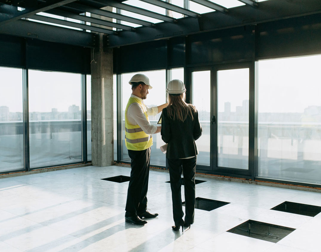 Zwei Personen mit Schutzhelmen inspizieren einen unfertigen Raum mit großen Fenstern und offenen Bodenplatten.
