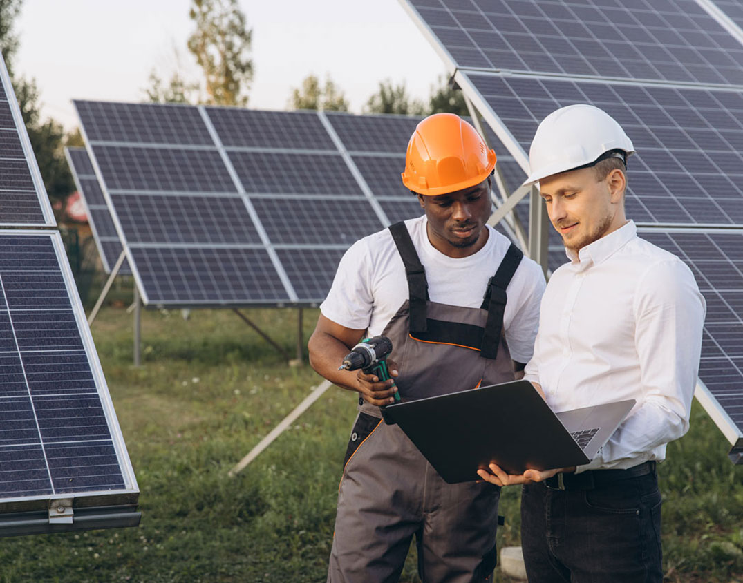 Zwei Arbeiter mit Schutzhelmen stehen zwischen Solarzellen im Freien.