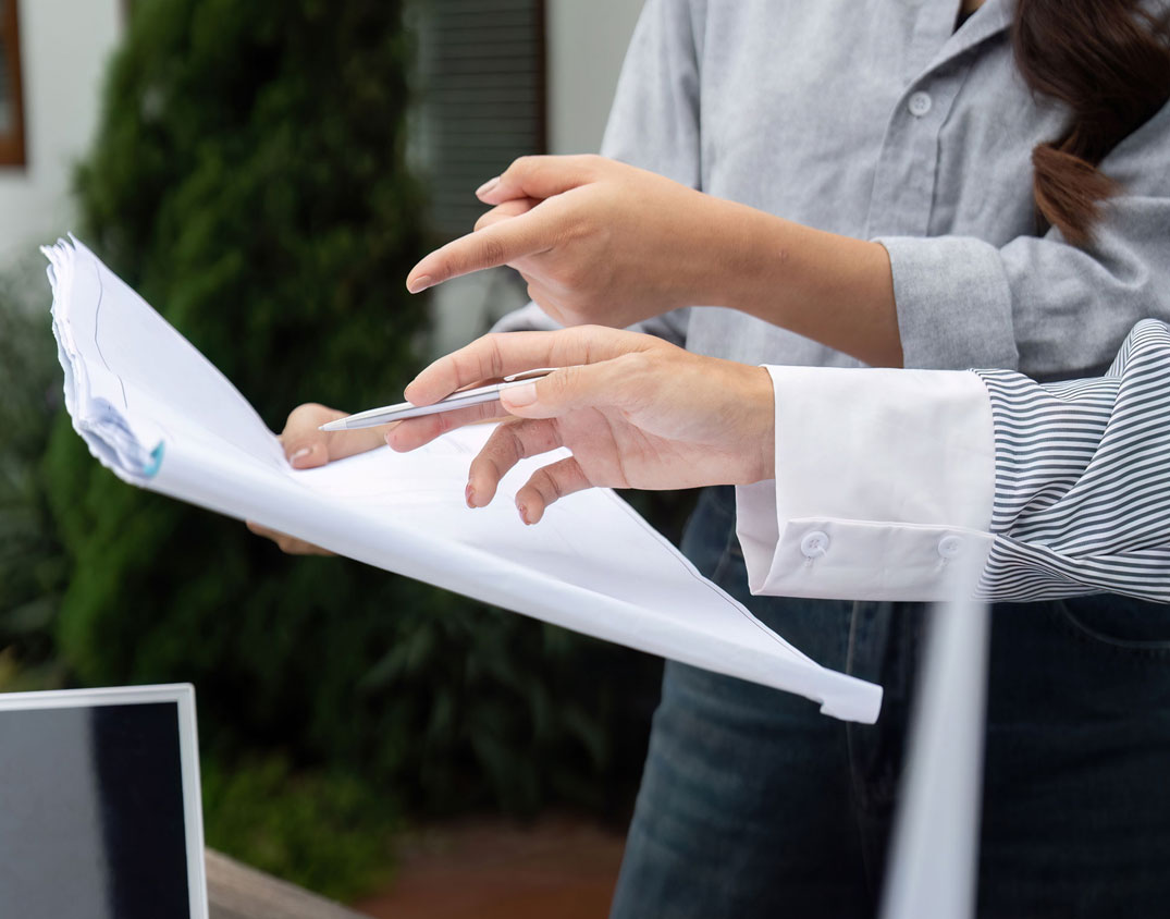 Zwei Personen prüfen Dokumente, wobei die eine auf etwas zeigt und die andere einen Stift in der Hand hält.